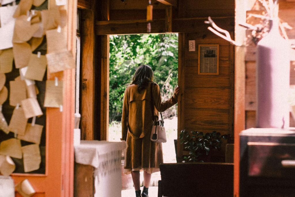 A woman in a warm coat steps out of a cozy cabin into the sunlight.