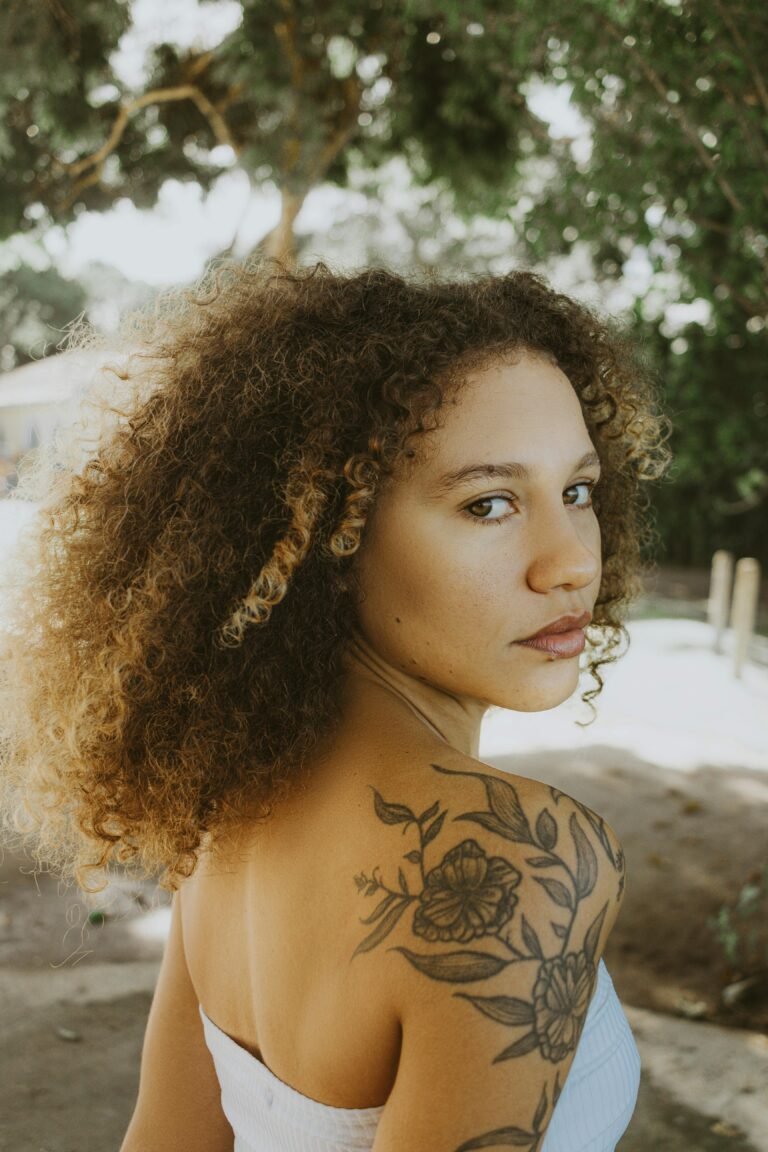A young woman with curly hair and tattoos looks over her shoulder in an outdoor setting.